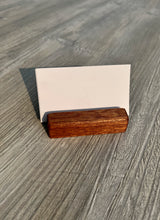 A rectangular wooden business card holder with a warm brown wood grain finish, holding a blank white card, set on a gray wood-textured surface.