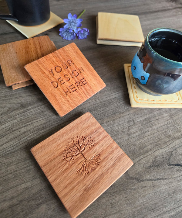 Square wooden coasters displayed on a dark wood tabletop, with one coaster showing the engraved text 'YOUR DESIGN HERE' and another showing an engraved tree motif. Ceramic cups and a small purple flower are placed around them as styling props.