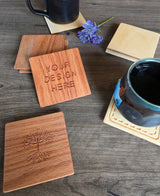 Square wooden coasters arranged on a dark wood tabletop, with two coasters showing engraved designs: one with the words 'YOUR DESIGN HERE' and one with a tree engraving. Ceramic mugs and a small purple flower are placed around them as styling props.