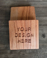 Two wooden coasters stacked, featuring the engraved text 'YOUR DESIGN HERE' on the front, photographed on a dark wood surface.
