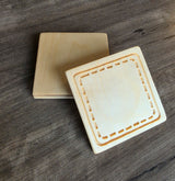 Three square wooden coasters with a dashed border design, stacked on a dark gray wood-textured surface.