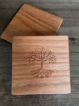 Two square wooden coasters with rounded corners, one in front of the other on a dark wood surface. The front coaster features an engraved tree with roots and branches.