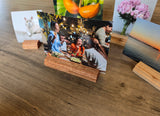 A wooden photo stand holding a printed photograph of a family dinner, displayed on a wooden tabletop alongside other sample prints and small wooden blocks.