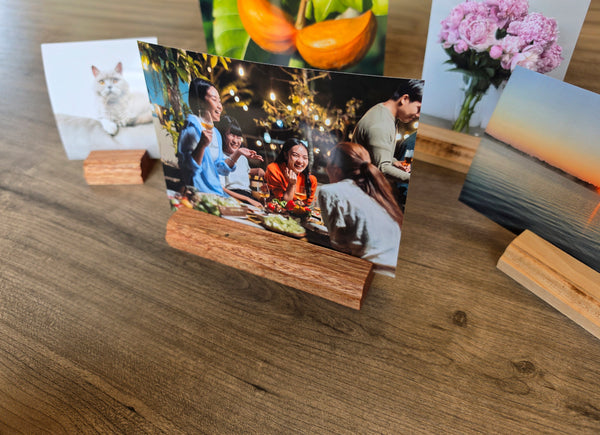 A wooden photo stand holding a printed photograph of a family dinner, displayed on a wooden tabletop alongside other sample prints and small wooden blocks.
