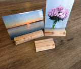 Three rectangular wooden photo stands arranged on a brown wood tabletop, holding printed photos of a sunset over water and pink flowers in a vase.
