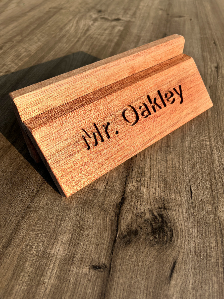 A personalized wooden desk nameplate engraved with 'Mr. Oakley', made from warm medium-brown wood and resting on a rustic dark wood tabletop.