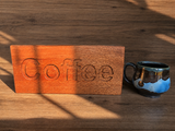 A rectangular wooden sign carved with the word 'Coffee' in a warm orange-brown wood tone, placed on a matching wooden surface next to a ceramic mug with blue, black, and brown glaze. Strong diagonal shadows are cast across the scene.