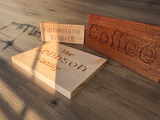 Three engraved wooden signs in different wood tones arranged on a wood-grain surface. One light natural wood plaque reads 'The Johnson Family', one medium brown plaque reads 'Coffee', and one darker brown plaque reads 'Ephesians 3:16-18'.