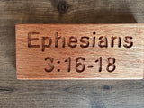 A rectangular wooden plaque engraved with the text 'Ephesians 3:16-18', displayed on a rustic dark wood background.