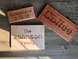Three engraved wooden sign plaques displayed on a dark wood tabletop. One plaque has a light natural wood finish and is engraved with 'The Johnson Family'.