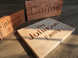 Three engraved wooden sign plaques displayed on a dark wood surface. The foreground plaque is light natural wood and engraved with 'The Johnson Family'.