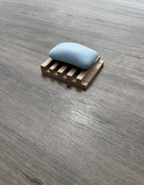A light blue soap bar resting on a small wooden soap dish, placed on a gray wood-textured surface.