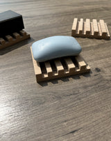 A light blue bar of soap resting on a wooden slatted soap dish, placed on a gray-brown wood tabletop. Two more wooden soap dishes are partially visible in the background.