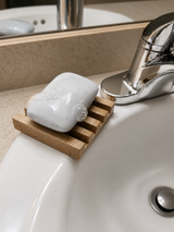 A white rectangular bar of soap resting on a small wooden soap dish placed on the edge of a white bathroom sink, with a chrome faucet visible.