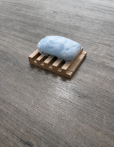 A light blue bar of soap covered in white lather, resting on a small rectangular wooden soap dish with slotted sides, placed on a gray wood-textured surface.